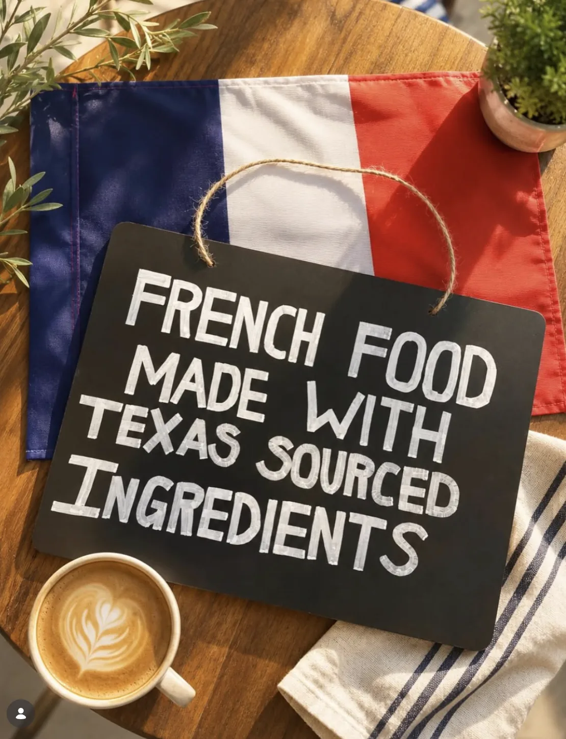 A chalkboard reading “French food made with Texas sourced ingredients”, resting on a French flag beside a latte and a blue-striped tea towel.