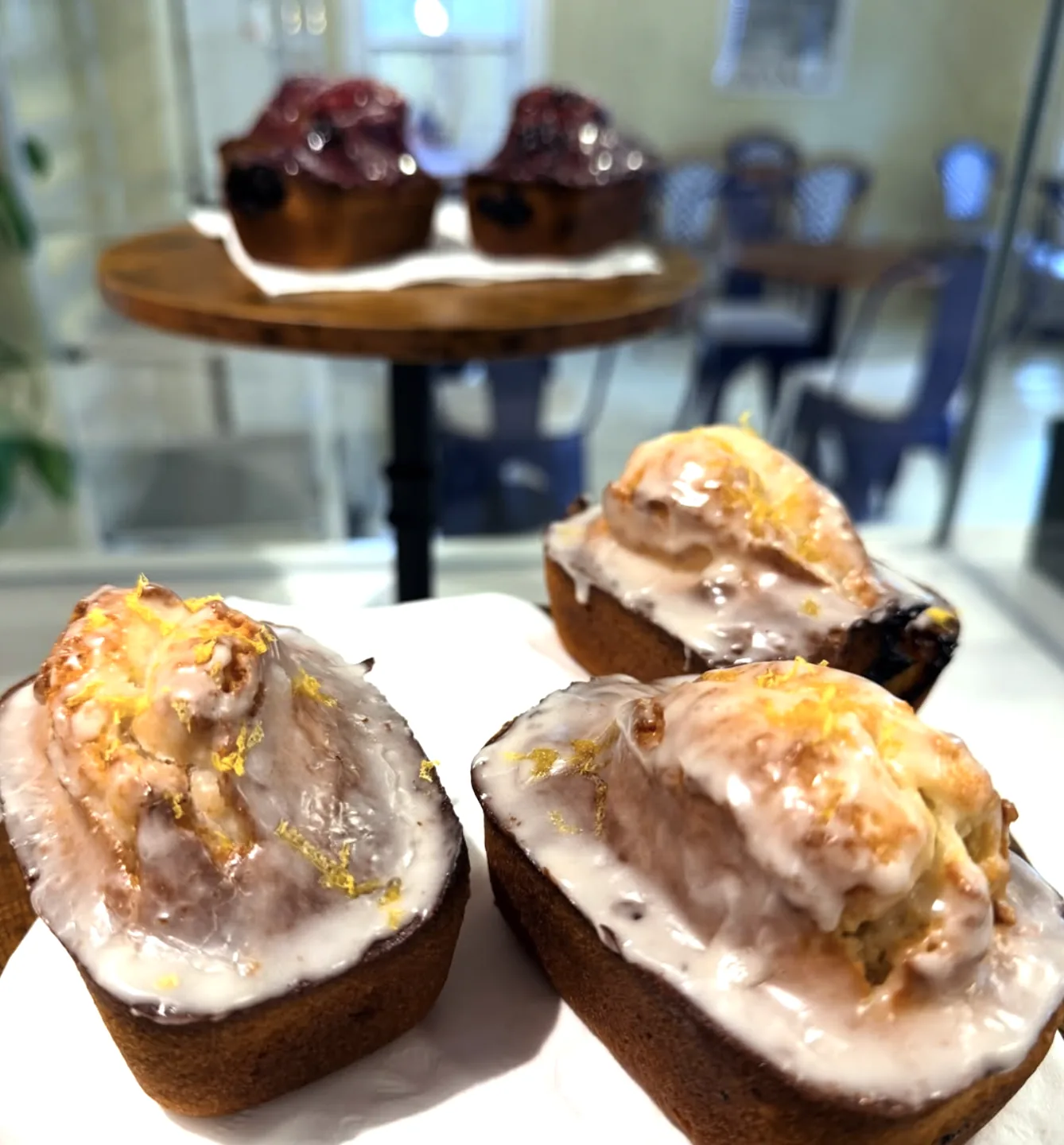 Lemon-glazed cakes with zest on top, with berry tarts on a wooden stand behind them inside the café.