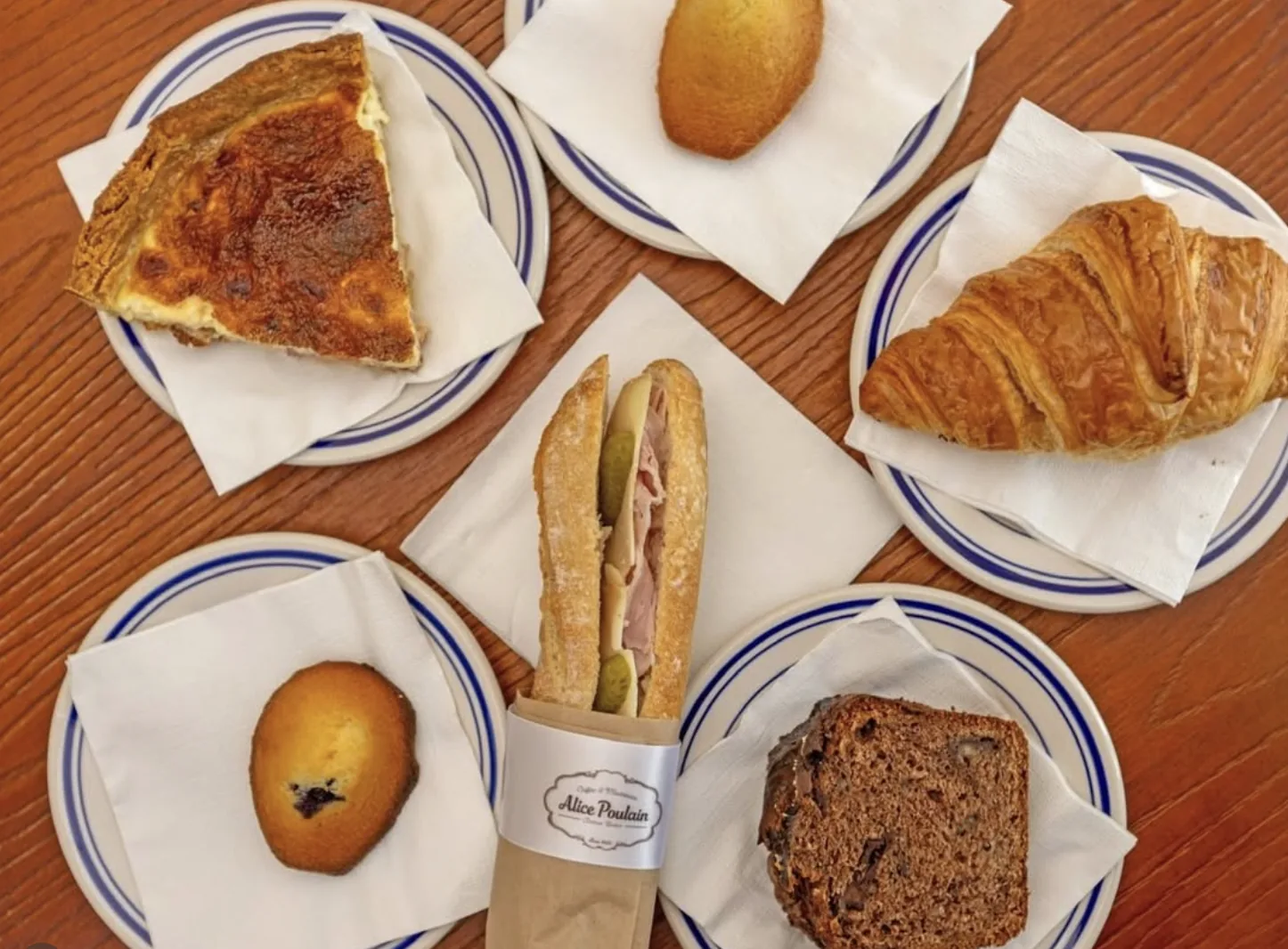 Overhead view of a quiche slice, madeleines, a butter croissant, a chocolate cake slice and a ham-and-cheese demi-baguette in Alice Poulain paper, each on blue-and-white striped plates on a wooden table.